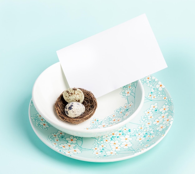 Table setting with Easter quail eggs in nest white blank sheet on floral pattern plates on blue