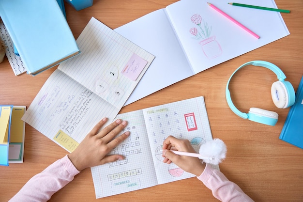 Table top closeup view of kid student hands writing doing math homework exercises in notebook with book on table Learning classes for elementary school pupils Study at home classroom concept