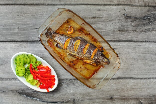 Table top view on grilled trout with lemons in glass baking pan, and bowl of green cucumber, onion and red pepper, on gray wood desk.