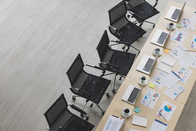 Table top view of wooden conference table with charts and graphs papers and laptop on it and six chairs lying in straight line beside the table in office. Concept for business meeting.