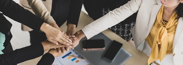 Table top view of a wooden conference table with charts and graphs papers on the table and six business women's hands put together cheering. Concept for business meeting.