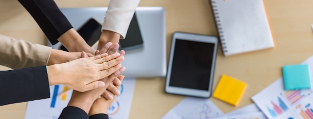 Table top view of a wooden conference table with charts and graphs papers on the table and six business women's hands put together cheering. Concept for business meeting.