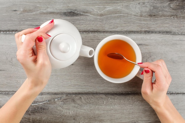 Table top view on young woman hands holding white ceramic teapot in one and silver spoon above cup of hot tea in other.