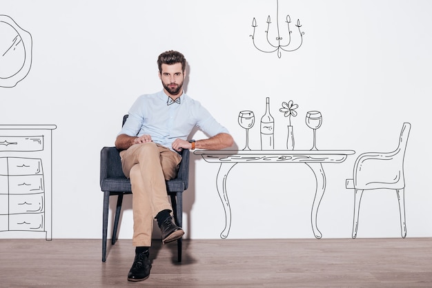 Table for two. Young handsome man keeping legs crossed and looking at camera while sitting in the chair against illustration of dining table in the background