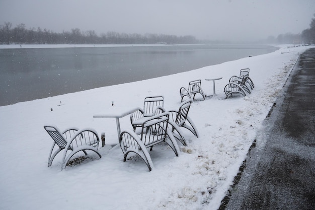 Tables and chairs under snow in cafe by the lake during the winter day