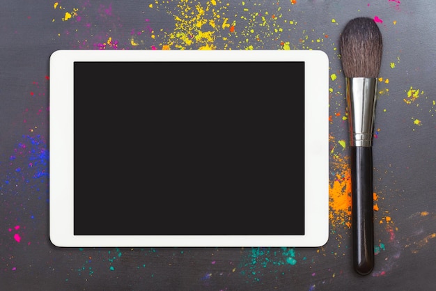 Photo tablet computer with blank screen with makeup brush on black background with colorful cosmetic powde