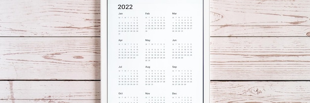 木の板の背景に2022年のカレンダーのオープンアプリとタブレットコンピューター。コンセプトビジネスまたはテクノロジーを使用してリストの目標を実行します。上面図、フラットレイ。バナー