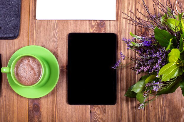 A tablet next to a cup of coffee and a notepad with purple flowers on it