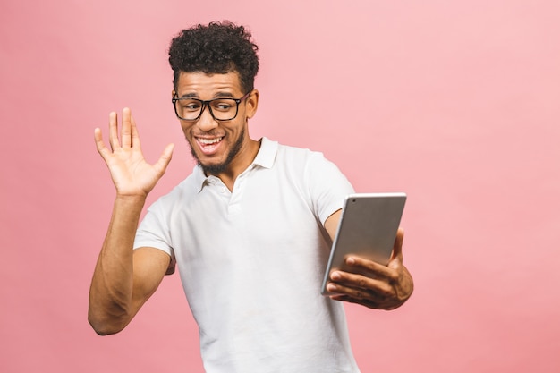 Tablet gebruiken. jonge knappe afrikaanse mens die holdingstablet glimlachen en spelen of een boeking app gebruiken die tegen roze muur wordt geïsoleerd.