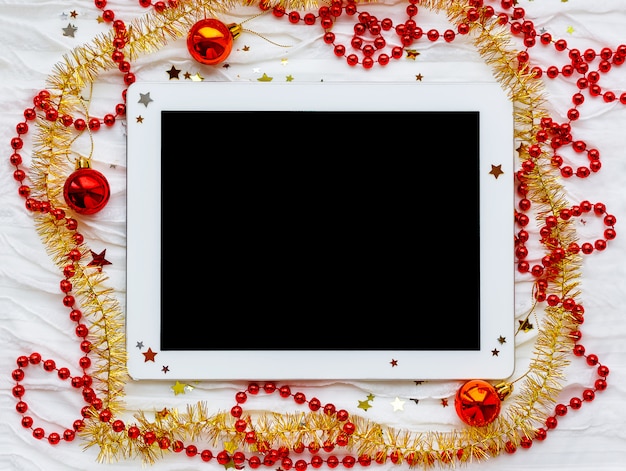 Tablet on winter holiday background. Christmas decorations - red and yellow tinsel, star sparkles, beads.