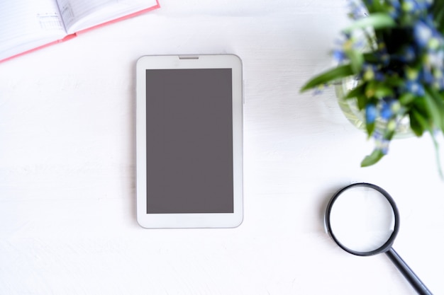 Tablet with black empty screen. Notebook, magnifier and flowers on table