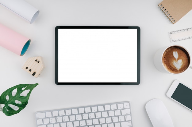 Tablet with blank and white screen on desk including a wooden mini house. keyboard, mouse, smartphone, notebook