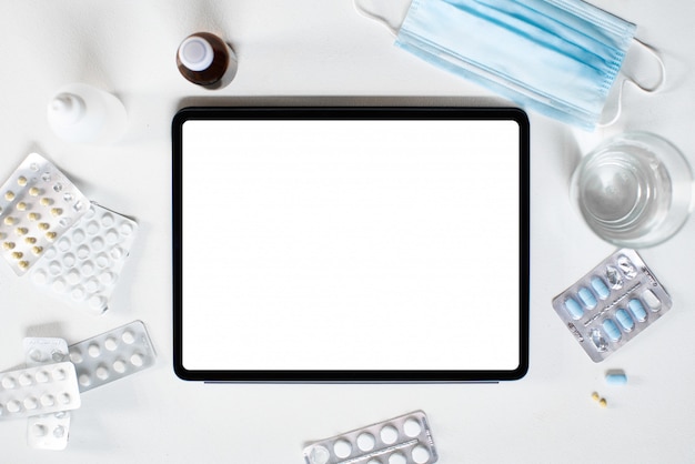 A tablet with an empty screen is lying on the table among medications medical supplies tablets and a surgical mask.