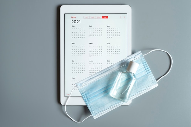 A tablet with an open calendar for 2021 year and protective medical mask and hand sanitizer on a gray