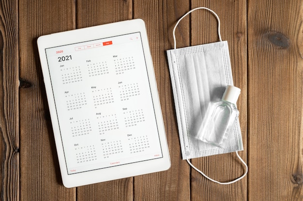 A tablet with an open calendar for 2021 year and protective medical mask and hand sanitizer on a wooden boards table background. covid-19 coronavirus protection concept in 2021