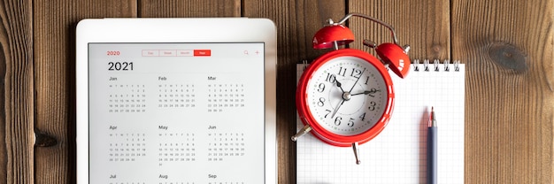 A tablet with an open calendar for 2021 year, a red alarm clock, chestnuts, and a spring notebook with a pen on a wooden boards table background. 