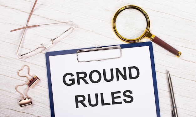 A tablet with the text ground rules on white paper a magnifier\
glasses golden paper clips and a pen lie on a white office table\
top view flat lay