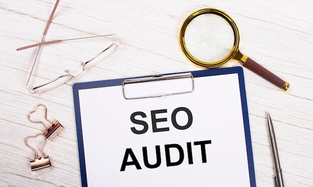 白い紙にSEO AUDITというテキストが付いたタブレット、拡大鏡メガネの金色のペーパークリップ、ペンが白いオフィスのテーブルに横たわっていますトップビューフラットレイ