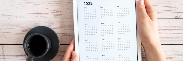 Tabletcomputer met een open app van kalender voor 2022 jaar en kopje thee of koffie in handen van de vrouw op houten achtergrond concept zaken of takenlijst doelen bovenaanzicht plat lag banner