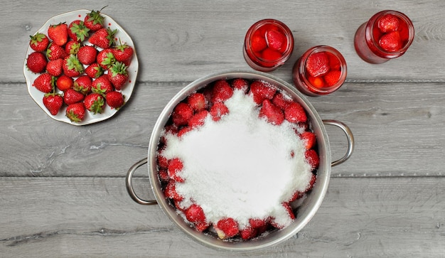 Tabletop photo - large steel pot of strawberries covered with crystal sugar, plate and glass bottles with more fruits around on gray wood desk.