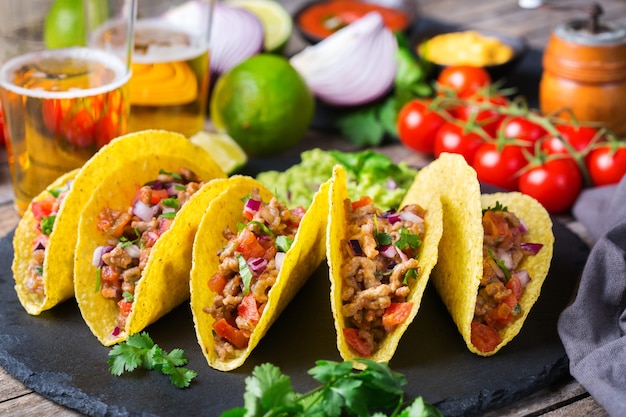 Taco shell ellow corn tortilla chips nachos with ground beef, mince, guacamole, red hot jalapeno chili salsa and cheese sauce with tequila or beer on a table.