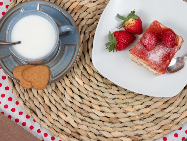 tafel met een plakje cake op een witte plaat met twee aardbeien en een lepel, en een blauwe beker met melk.