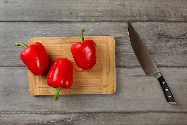 Tafelbladweergave, drie rode paprika's, besproeid met druppels water, klaar om te worden gesneden op een snijplank, met een koksmes aan de zijkant.