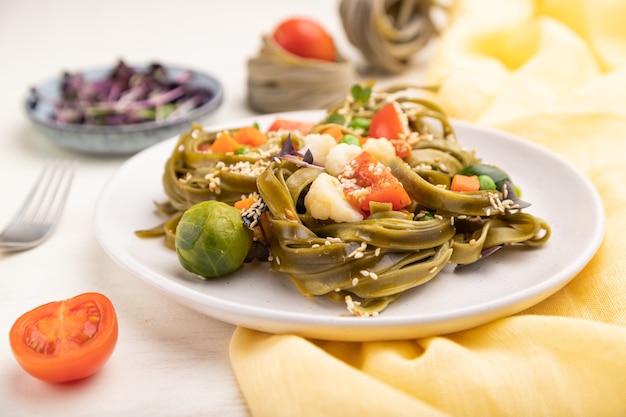 Tagliatelle di pasta verde agli spinaci con pomodoro, piselli e germogli microgreen su una superficie di legno bianca e tessuto giallo