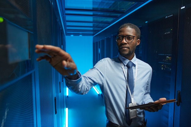 Taille omhoog portret van Afro-Amerikaanse man die naar de serverkast reikt terwijl hij met een supercomputer in het datacenter werkt en een tablet vasthoudt, kopieer ruimte