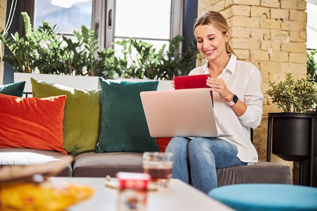 Taille omhoog portret van een gelukkige aantrekkelijke vrouw in jeans en wit overhemd die naar het scherm van een moderne laptop kijkt terwijl ze het cadeau in handen houdt