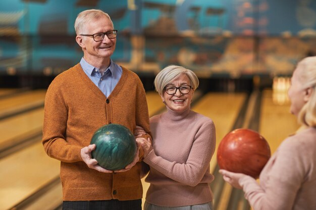 Taille omhoog portret van een groep gelukkige senioren die bowlingballen vasthouden en chatten terwijl ze genieten van actief entertainment op een bowlingbaan, kopieer ruimte