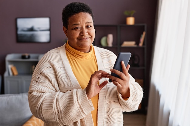 Taille omhoog portret van een zwarte oudere vrouw die een smartphone vasthoudt die thuis bij het raam staat en ernaar kijkt