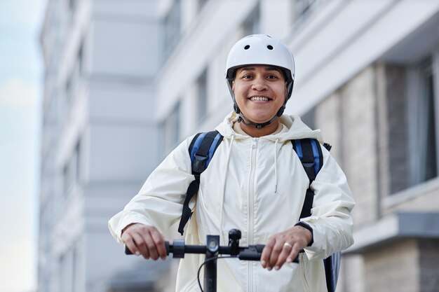 Taille omhoog portret van glimlachende voedselbezorger die een helm draagt terwijl hij op een elektrische scooter rijdt in ci