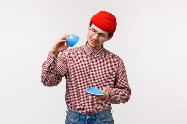 Taille-omhoog portret van grappige Europese man met snor in hipster beanie, bril, kijk verdrietig als er geen koffie meer over is, kijk somber naar lege beker, wil deze heerlijke thee nog wat drinken,