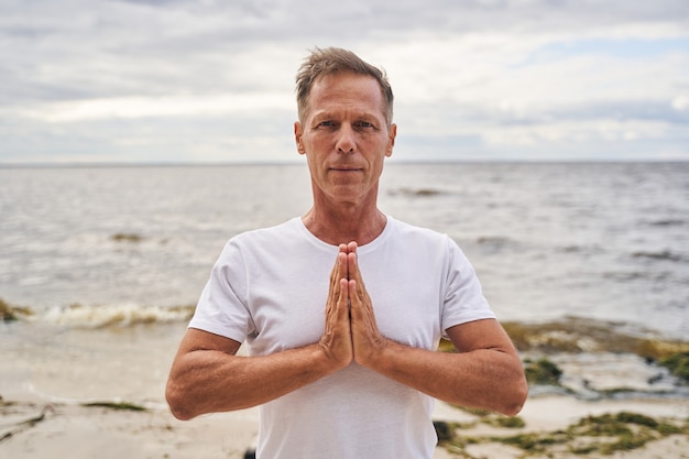 Taille omhoog portret van sportief mannetje dat aan de kust staat en handen vasthoudt in namaste-positie