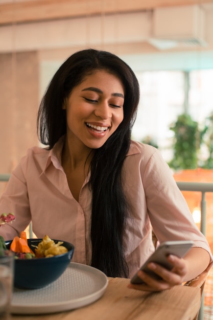 Taille omhoog van een gelukkige jonge vrouw die lacht met haar mond open en naar het scherm van haar smartphone kijkt terwijl ze eet in een café