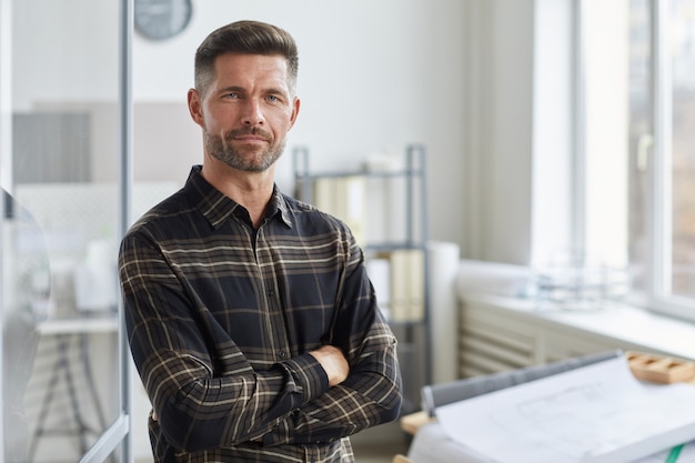 Taille portret van zelfverzekerde bebaarde architect staand met armen gekruist door tekentafel in kantoor,