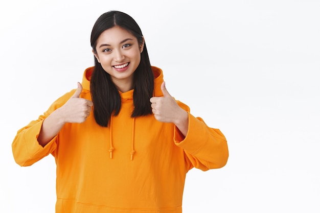 Taille-up portret aziatisch millennial meisje in oranje hoodie met duim omhoog in like of goedkeuring, accepterend glimlachen, positieve feedback geven, goed product beoordelen, staande witte muur