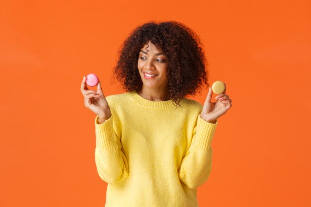 Taille-up portret dromerige schattige moderne Afro-Amerikaanse vrouw trakteert zichzelf, eet desserts op cheat-dag, slaat dieet over, houdt twee smakelijke macarons en glimlacht, zoals snoepjes, staat oranje