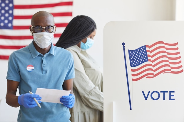 Taille-up portret van Afro-Amerikaanse man permanent door stemhokje versierd met vlag van de VS en op post-pandemische verkiezingsdag, kopie ruimte