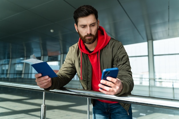 Taille-up portret van de jonge blanke man die in de luchthavenlounge staat en zijn tickets op de smartphone controleert. Reizen met het vliegtuigconcept