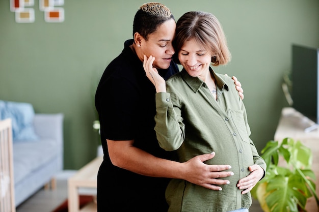 Taille-up portret van een gelukkig lesbisch koppel dat een baby verwacht en liefdevol omhelst in de politie in het interieur