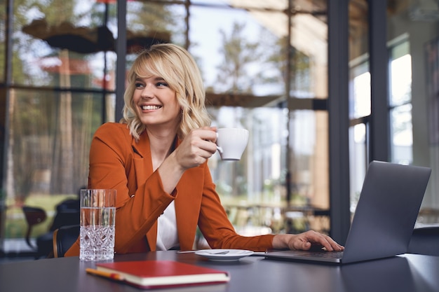 Taille-up portret van een lachende vrouwelijke ondernemer zittend met een koffiekopje aan tafel