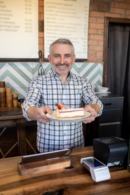 Taille-up portret van een vriendelijke bakkerijmedewerker die een stuk slagroomtaart overhandigt aan zijn klant