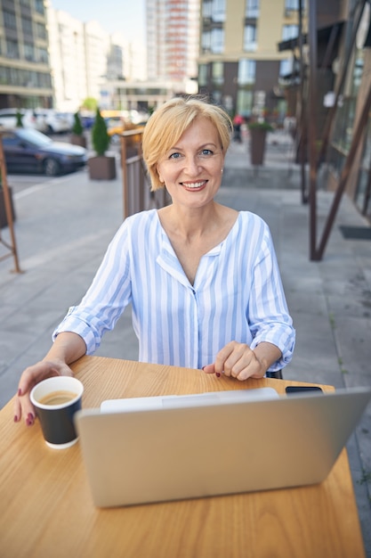 Taille-up portret van een vrolijke blonde zakenvrouw die geniet van haar koffiepauze in een straatcafé
