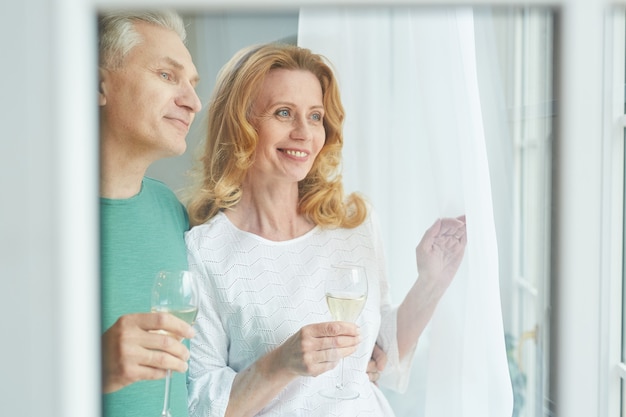 Taille-up portret van elegante volwassen paar genieten van champagne terwijl u uit het raam kijkt, schot van achter glas, kopie ruimte