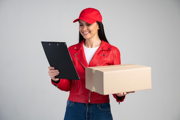 Taille-up portret van gelukkige vrouwelijke koerier in rood uniform met kartonnen pakket en kijkend naar de map met gelukkige emoties. Indoor studio-opname geïsoleerd op een grijze achtergrond