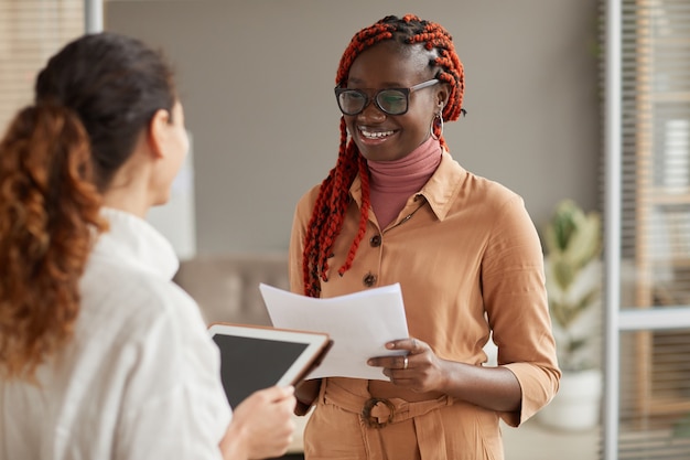 Taille-up portret van jonge afro-amerikaanse zakenvrouw praten met vrouwelijke manager en vrolijk glimlachen terwijl staande in moderne kantoor interieur