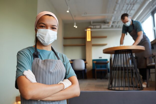 Taille-up portret van jonge serveerster met masker en schort kijkend naar camera terwijl hij in café staat met man die tafels op de achtergrond schoonmaakt, covid-veiligheidsmaatregelen, kopieer ruimte