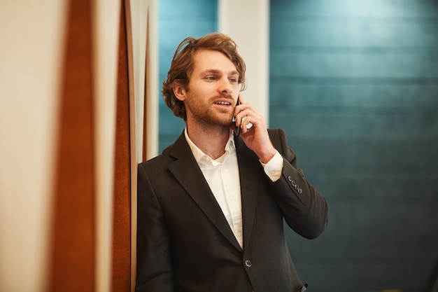 Taille-up portret van knappe bebaarde zakenman spreken door smartphone en wegkijken terwijl leunend op de muur in minimaal kantoor interieur, kopie ruimte